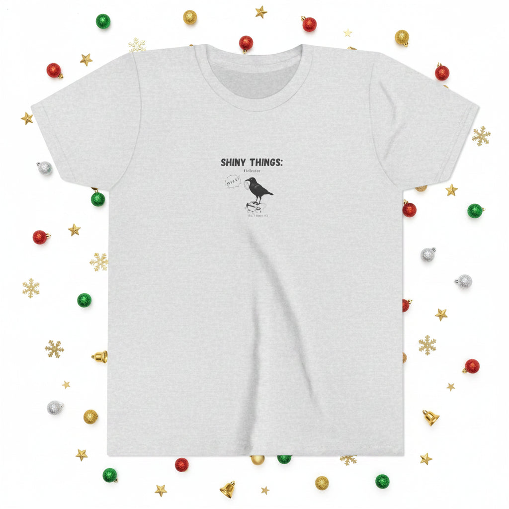 Ash grey t-shirt with graphic of a black crow and speech bubble saying MINE. The headline is shiny things. The background cartoon shows shiny object the crow collected.
