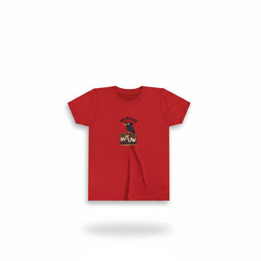 A red short-sleeve T-shirt with a graphic print of a black bird and the words 'BACKYARD OUTLAW' is displayed in front of a wooden fence and a wanted poster. A large black bird with spread wings perches on the fence behind the shirt, and a warm sunset sky fills the background. AI created from prompt by Malika Bourne Legacy of Ray the tlaking crow.