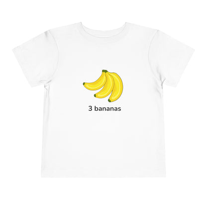 A white short-sleeve crewneck t-shirt featuring a graphic of three yellow bananas with the text '3 bananas' printed underneath, displayed on a clean white background. Toddler “3 bananas” tee in white – fruit counting design