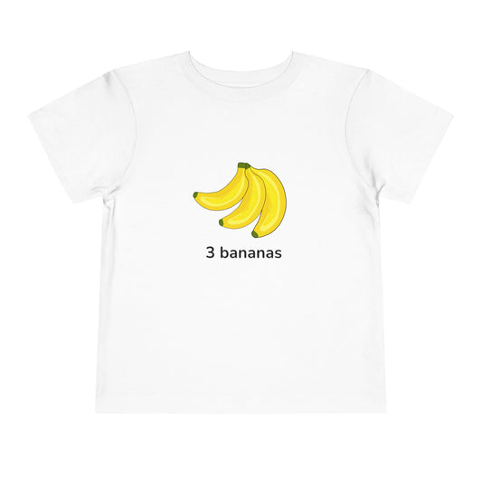 A white short-sleeve crewneck t-shirt featuring a graphic of three yellow bananas with the text '3 bananas' printed underneath, displayed on a clean white background. Toddler “3 bananas” tee in white – fruit counting design