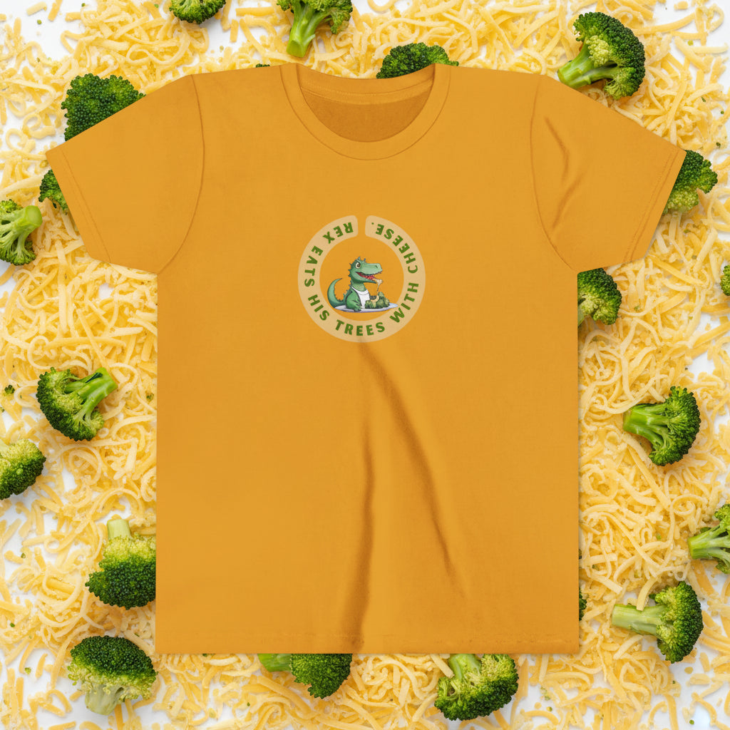 A mustard yellow short-sleeve t-shirt with a circular graphic on the chest featuring a green cartoon dinosaur and the text 'Rex eats his trees with cheese. The backgorudn is grated cheese and broccoli florettes