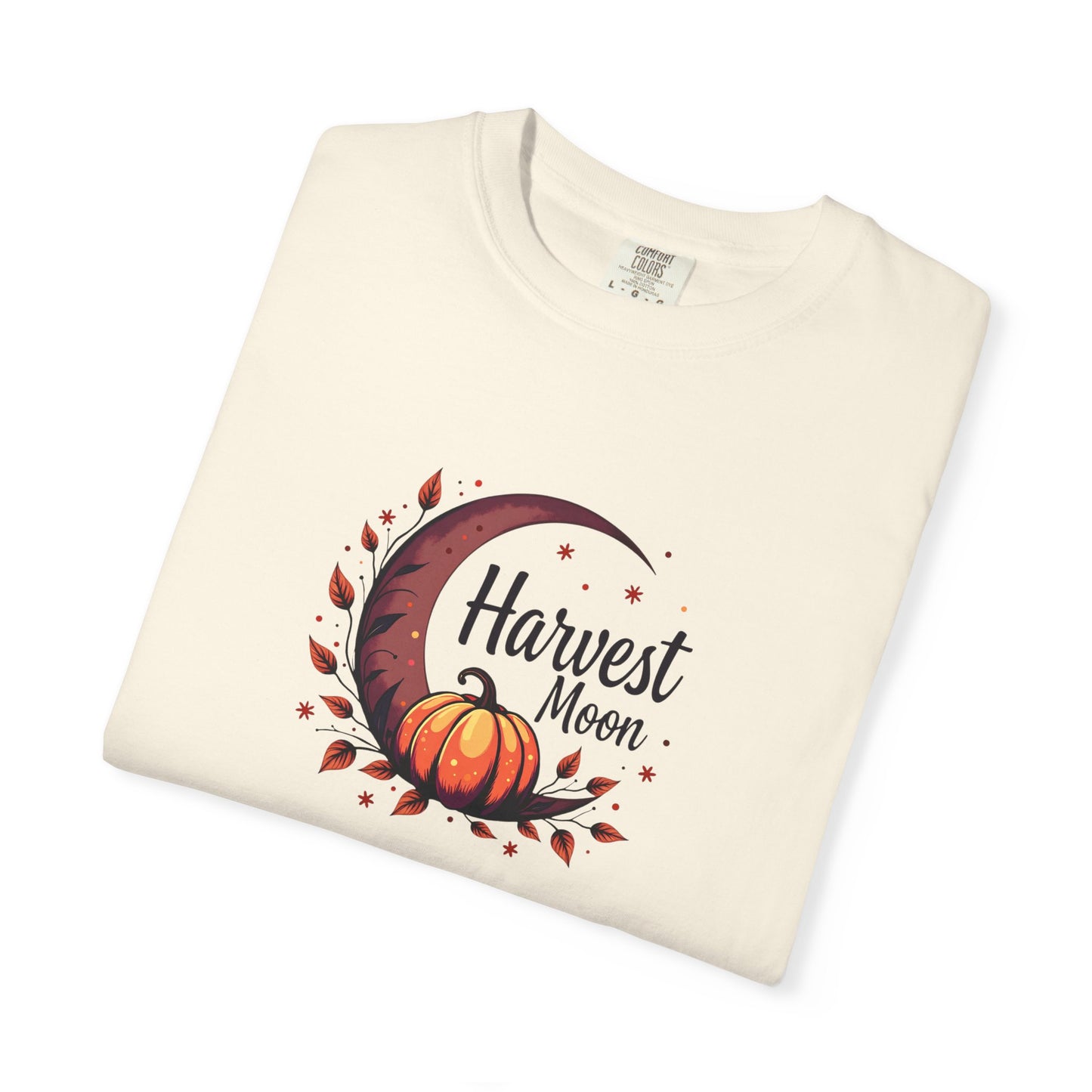A folded ivory/ cream-colored t-shirt featuring a graphic print of an orange pumpkin, a dark brown crescent moon, and autumn leaves with the text 'Harvest Moon' on a white background.