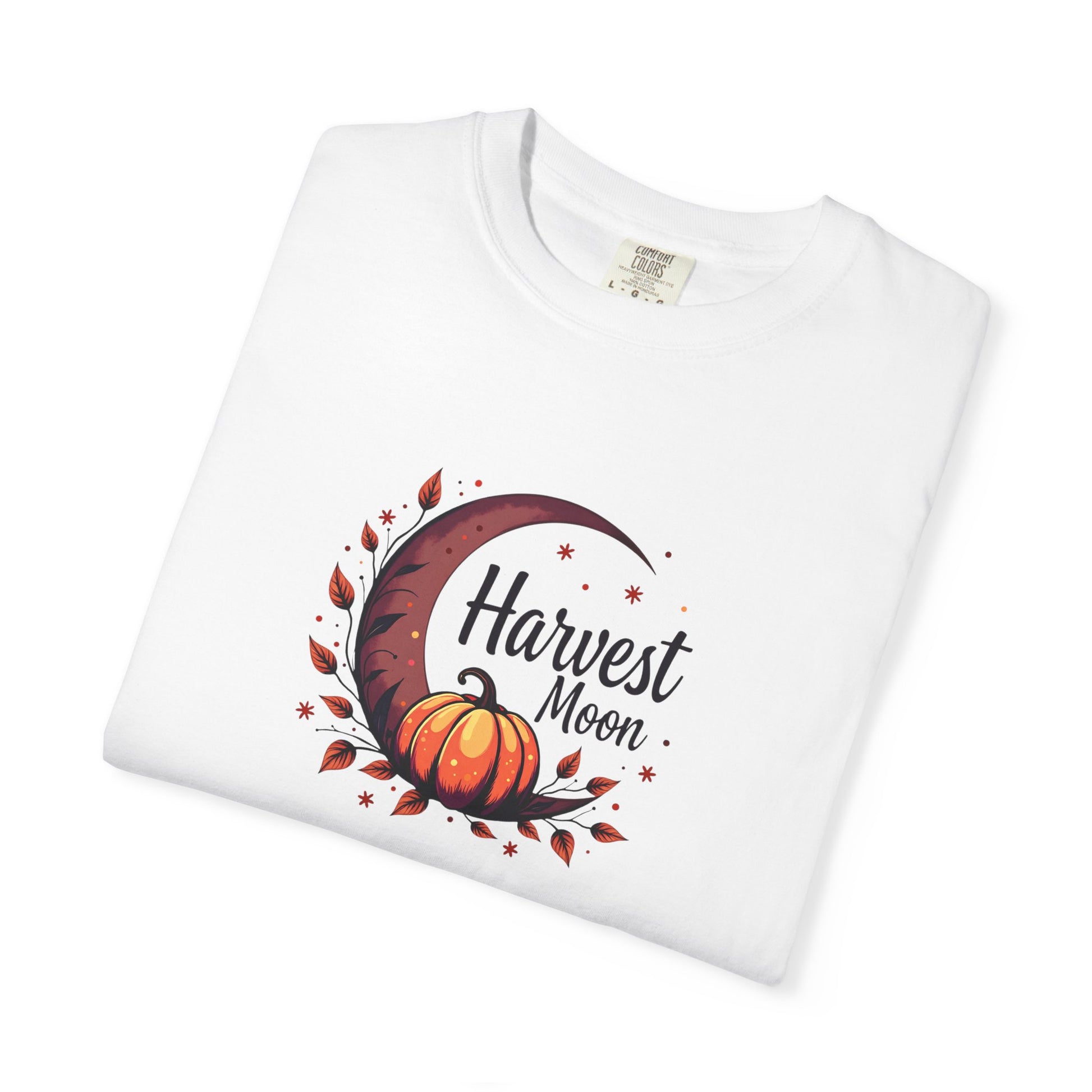 A folded white crewneck t-shirt featuring a graphic print of an orange pumpkin, a dark brown crescent moon, and autumn leaves with the text 'Harvest Moon'.