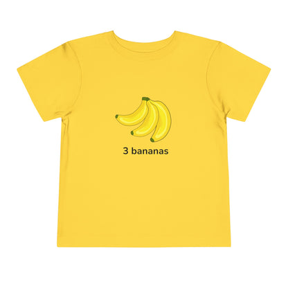 A yellow short-sleeve t-shirt with a graphic of three bananas and the text '3 bananas' printed on the chest, displayed on a solid white background. Toddler “3 bananas” T‑shirt in yellow – fruit counting design