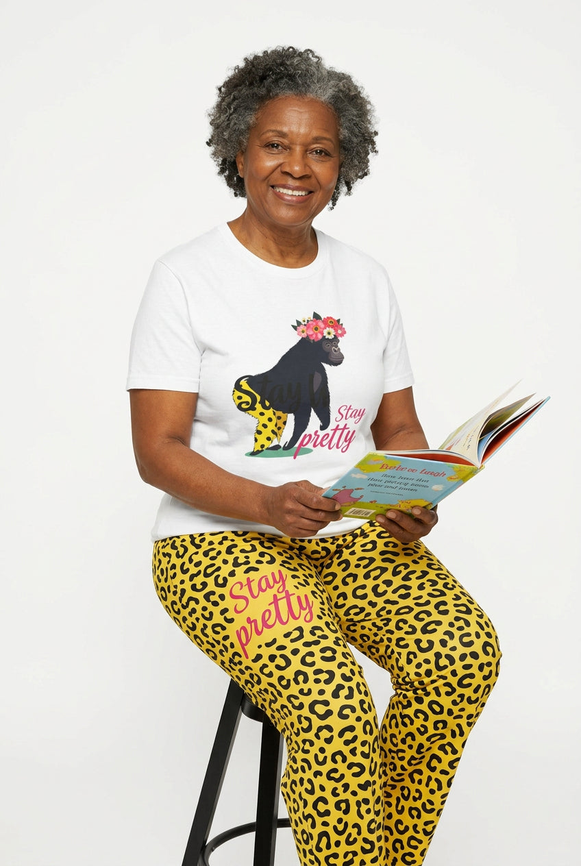 AI model is an older, dark-skinned woman wearing this shirt.“Girl‑Girl™ gorilla wearing a flower crown and patterned shorts on a white unisex tee with the phrase ‘Stay Pretty.’” AI model wears yellow cheetah-print pants. She appears to read a children's book.