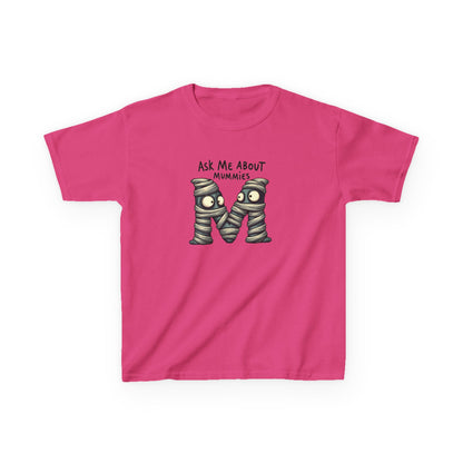 A bright pink. heliconia short-sleeve t-shirt with a graphic of the letter 'M' styled as two mummies wrapped in bandages, with the text 'ASK ME ABOUT MUMMIES' above it, shown on a plain white background.