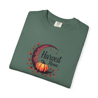 A folded sage green t-shirt with a 'Harvest Moon' graphic featuring an orange pumpkin, crescent moon, and autumn leaves, displayed on a clean white background.