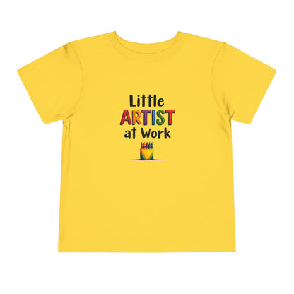A yellow short-sleeve toddler's t-shirt with a colorful graphic on the chest that reads 'Little ARTIST at Work' above a small crayon box illustration, shown on a plain white background.