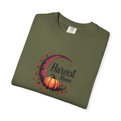 An olive green folded t-shirt with a 'Harvest Moon' graphic featuring an orange pumpkin, crescent moon, and autumn leaves on a white background.