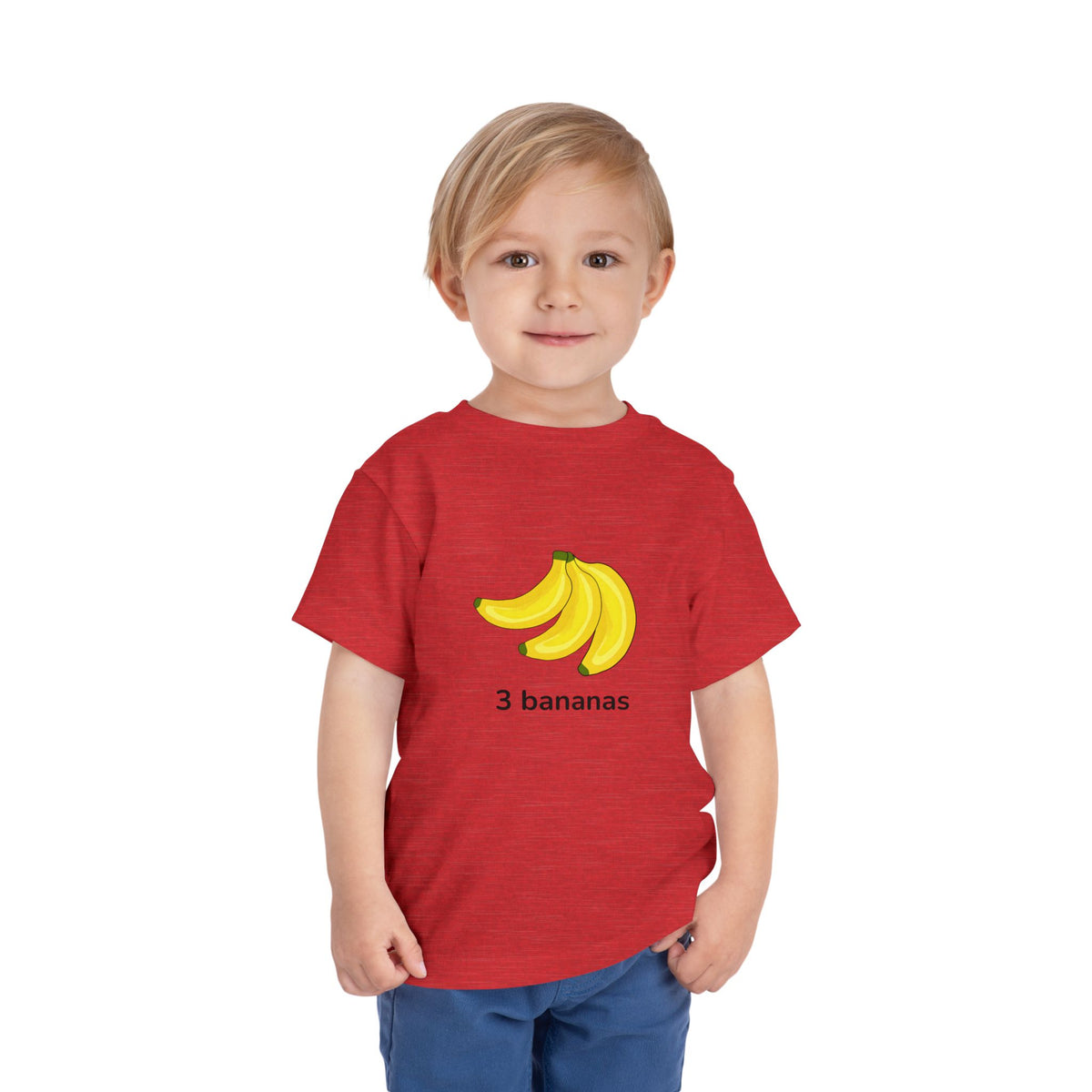 A young child wearing a red heathered t-shirt with a graphic of three yellow bananas and the text '3 bananas' printed on the front, paired with blue pants, set against a plain white background.