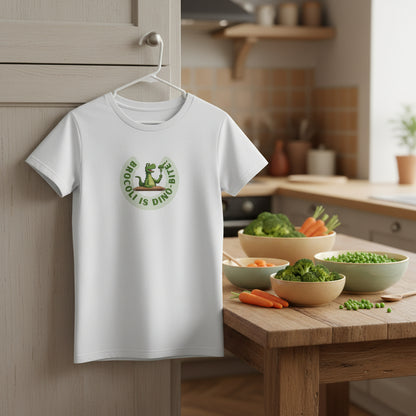 A white short-sleeve crewneck t-shirt featuring a circular green graphic on the chest with a cartoon dinosaur and the text Broccoli is dino-bite. Colorful background