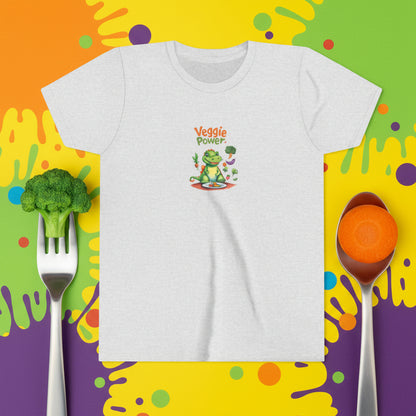 A light heather gray short-sleeve t-shirt with a colorful graphic on the chest. The graphic features a cartoon green dinosaur or aligator holding a carrot and broccoli, sitting at a table with a plate of veggies. Shirt with the text 'Veggie Power' above it.